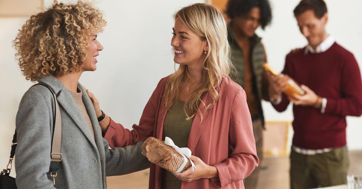 A blonde hostess smiling warmly while welcoming a friend into her home with a wrapped gift as arriving house guests gather in the background, illustrating thoughtful hospitality preparation.