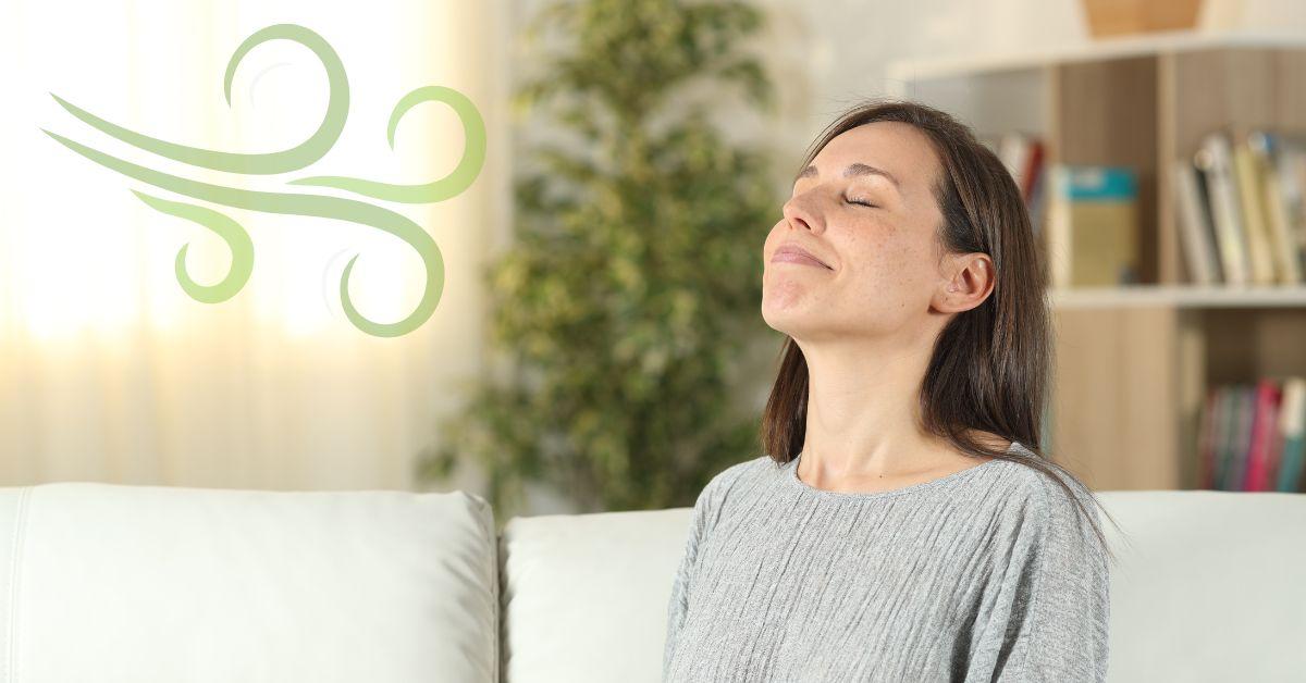 A relaxed woman sitting on a white living room sofa with her eyes closed, smiling as she breathes in deeply next to a green graphic icon of a fresh breeze, illustrating the benefits of pristine indoor air quality.