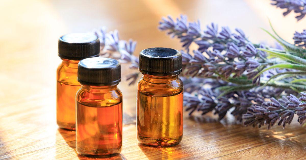 Three small amber glass apothecary bottles with black screw caps sitting on a wooden surface next to a bundle of dried purple lavender blossoms, illustrating the natural household applications of lavender essential oil.