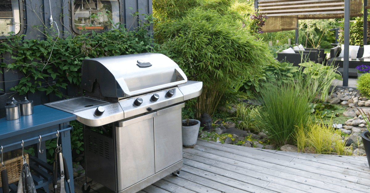 A polished, multi-burner stainless steel grill sitting on a wooden backyard deck next to a prep table, surrounded by lush green garden landscaping and an outdoor seating area, ready for summer family barbecues.