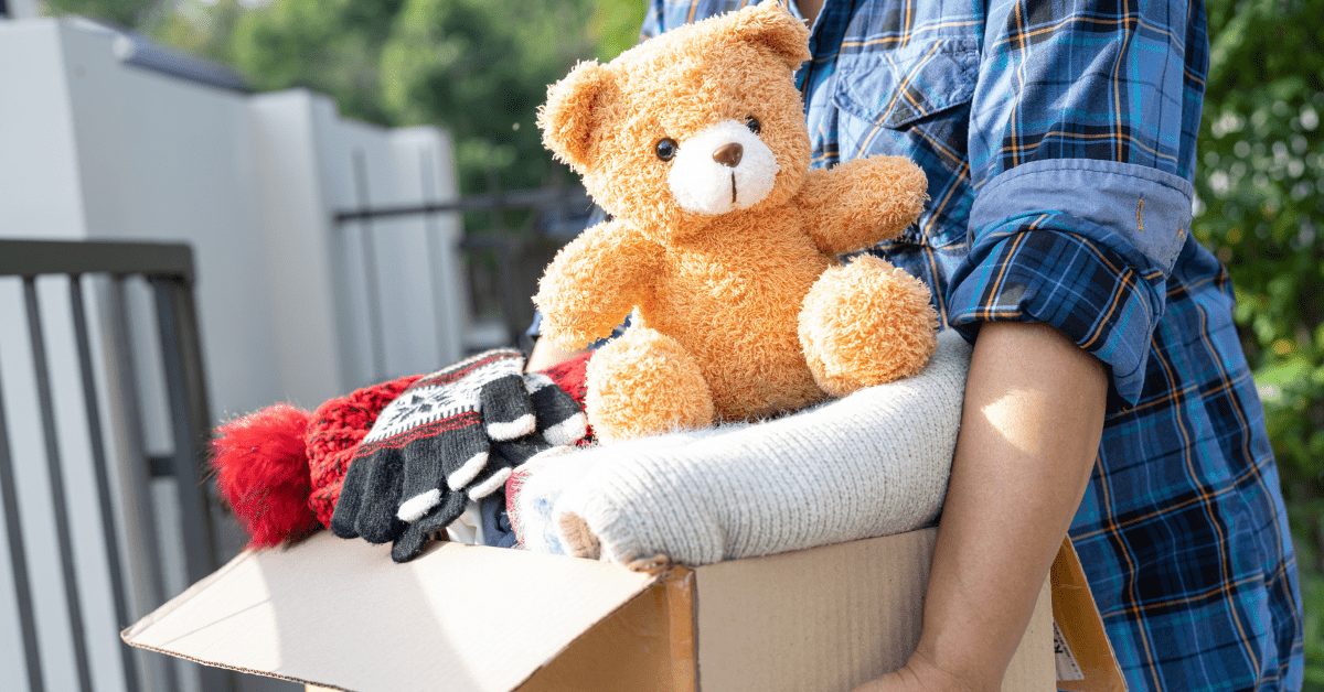 A close-up shot of a person wearing a blue plaid shirt holding a cardboard donation box filled with folded winter clothes, knit gloves, a warm hat, and a brown teddy bear, highlighting ways to support local long island charitable organizations.