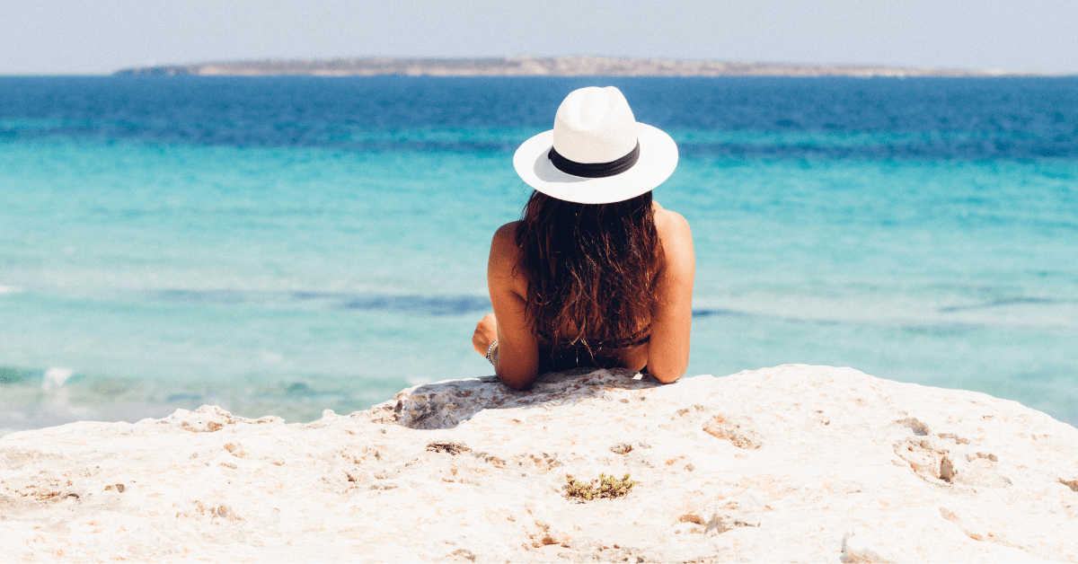 A woman wearing a white sun hat sits on a rocky shoreline looking out over turquoise ocean waters, illustrating the coastal beach lifestyle that requires proactive maintenance routines to keep sand out of your house.