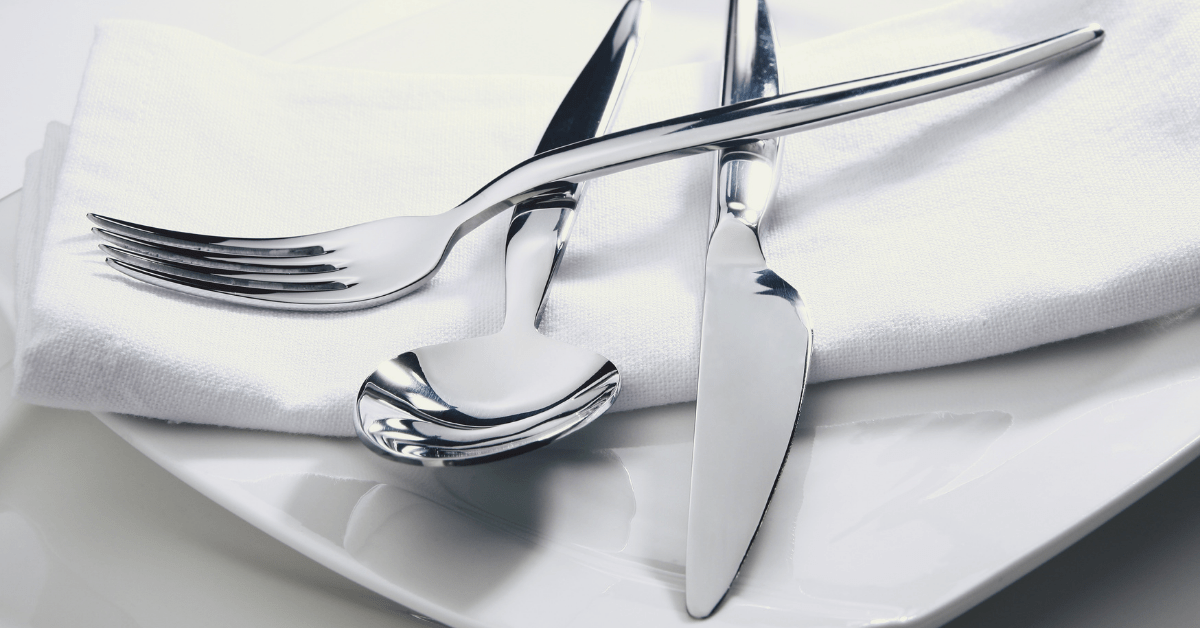 A close-up photograph of a gleaming, polished stainless steel fork, spoon, and knife arranged across a folded white cloth napkin on a dinner plate, illustrating how to clean silverware to a brilliant finish for formal holiday table settings.