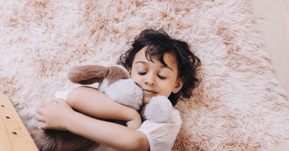 A young child lying on a soft, shag rug with closed eyes, happily hugging a favorite plush toy close to their face, emphasizing the importance of knowing how to clean a stuffed animal to safely remove germs, dust mites, and dirt from beloved playthings.