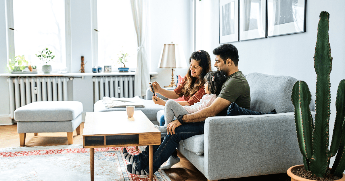 A happy family sitting together on a grey sofa in a bright, spotless living space, demonstrating the pristine results of taking the time to deep clean your living room for a healthier home environment.