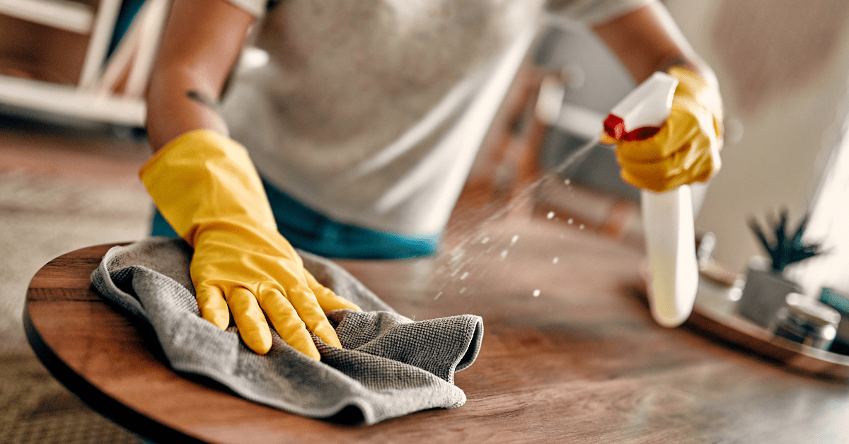 Person in yellow rubber gloves spraying and wiping a wood surface illustrating common house cleaning mistakes to avoid for a cleaner home